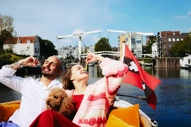 Laughing couple with a small brown dog lounge on orange cushions, Amsterdam flag and drawbridge behind