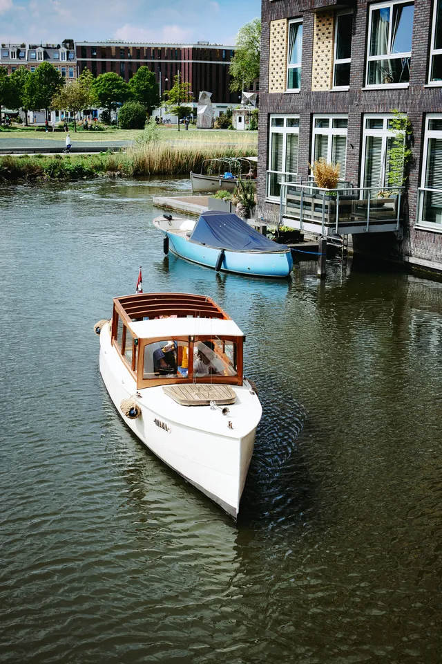 White and wood saloon boat Diana cruising an Amsterdam canal past a blue moored boat and brick apartments
