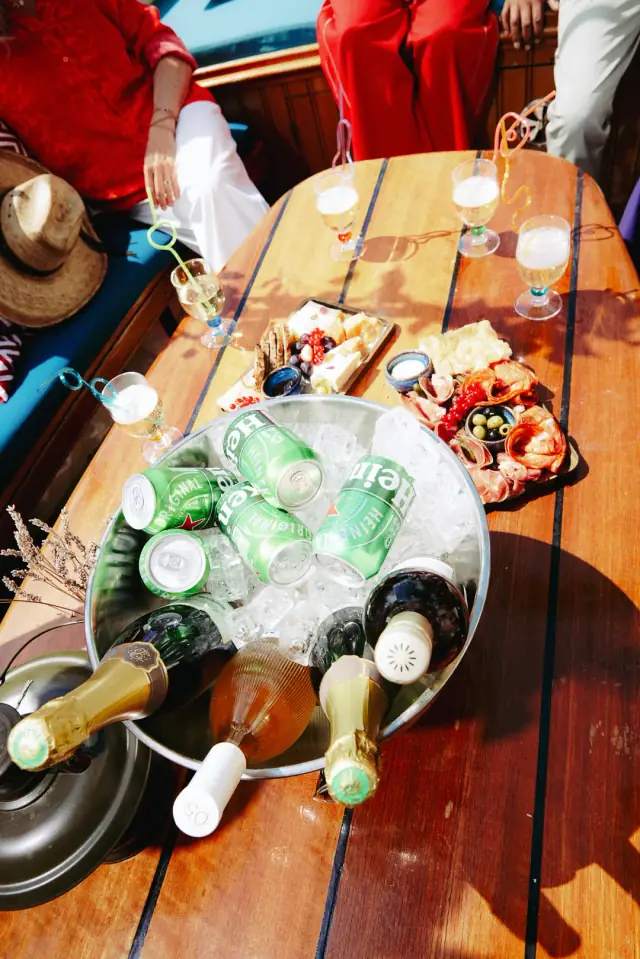 Ice bucket of beer cans and bottles beside glasses and charcuterie board on a wooden boat table