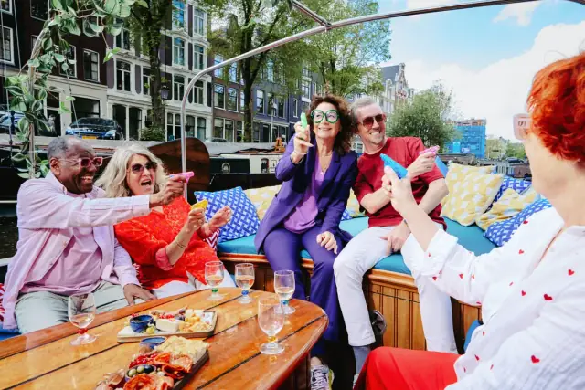 Group of laughing friends in bright clothes raise small party blowers over a wooden table with wine glasses and snack boards