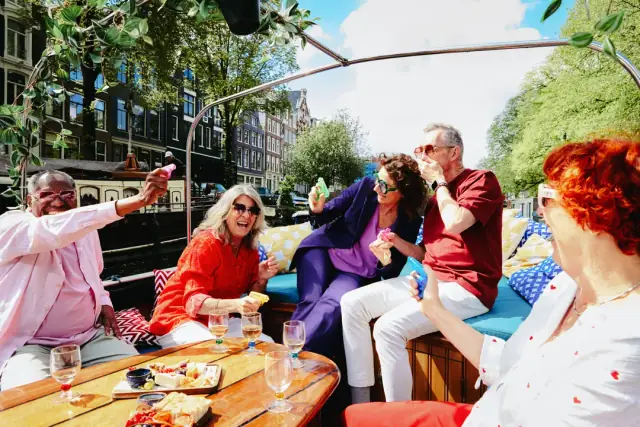 Five laughing friends in colourful clothes raise toys around a wooden table of snacks and wine glasses on a canal boat