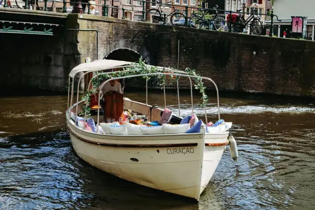 White canal boat Curaçao with a canopy and ivy garland, approaching a brick bridge arch in Amsterdam