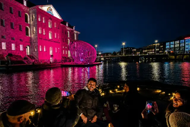 Smiling passengers photograph a pink-lit building and glowing lattice sphere from a boat at night, Amsterdam