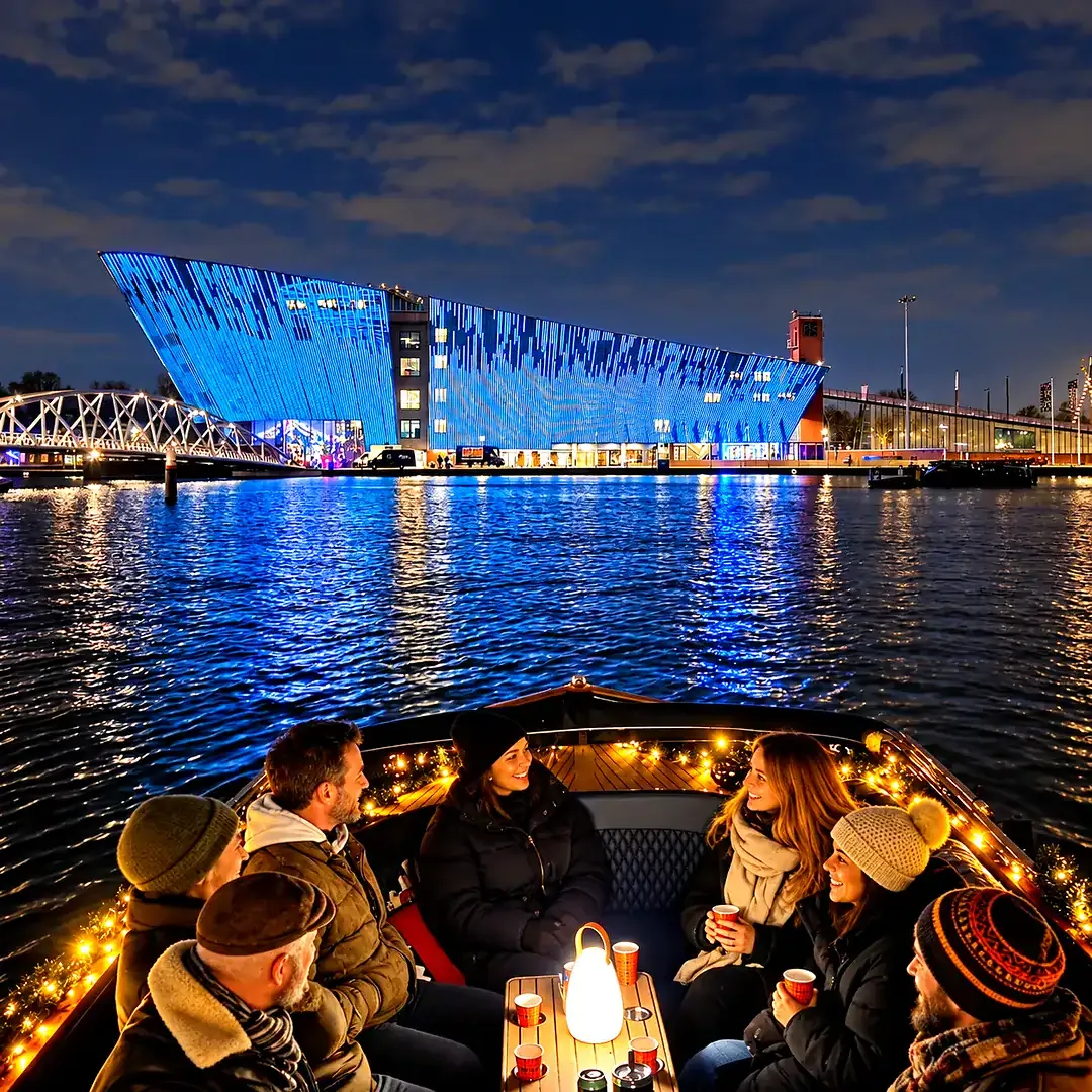 Guests with mulled wine cups on the boat at night, the lit NEMO building across the water
