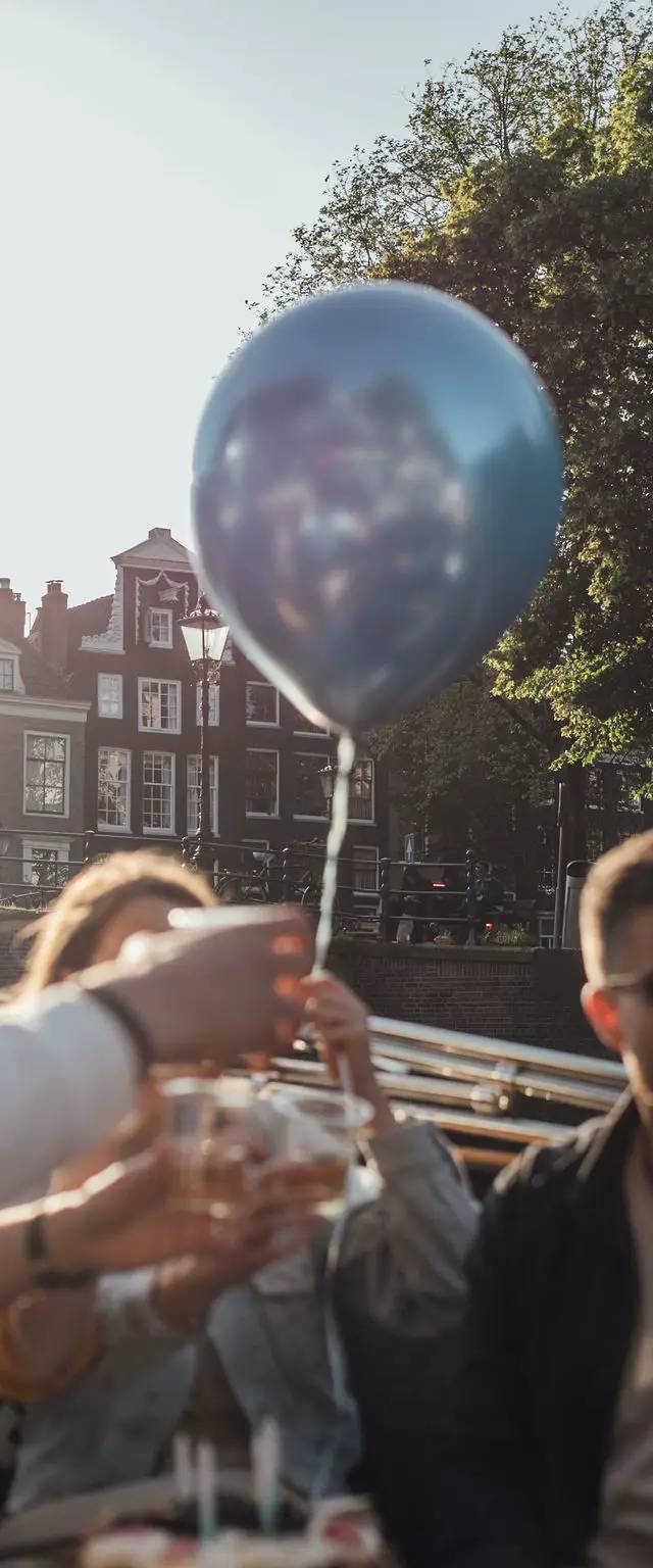 Friends raising glasses in a toast beneath a blue balloon, canal-side houses and trees behind