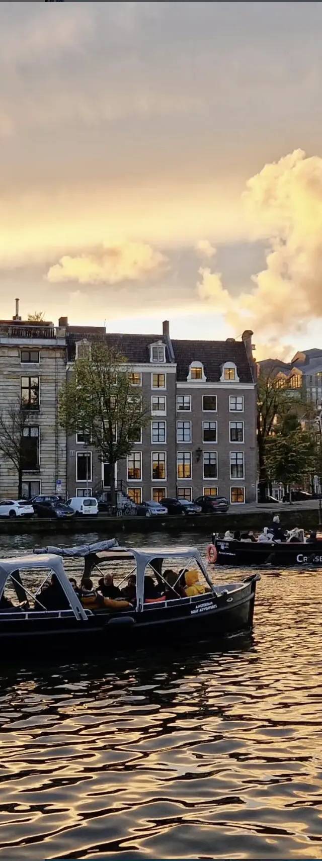 Open canopied boat with passengers on a golden-lit Amsterdam canal, canal houses and sunset clouds behind
