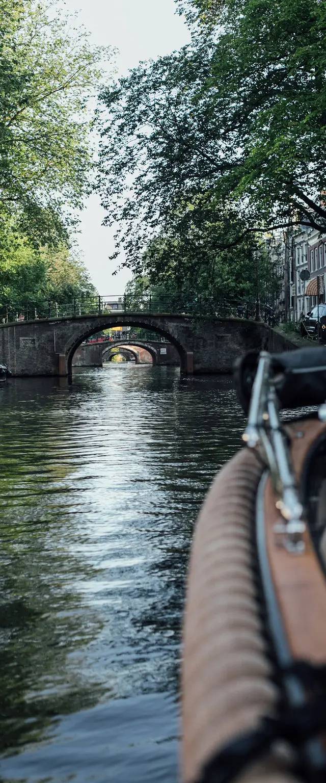 Stone arched bridges crossing a tree-lined Amsterdam canal, seen from the bow of a boat