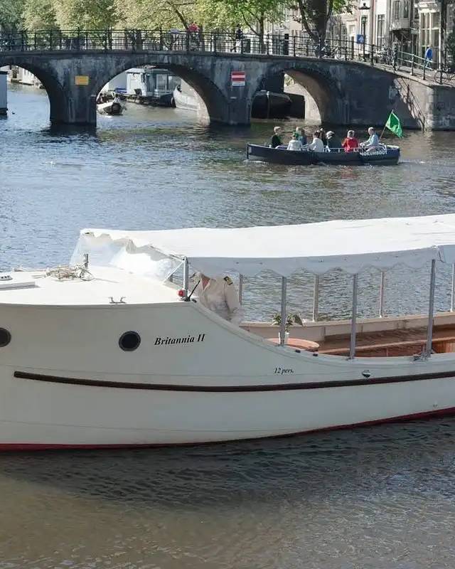 White canal boat Britannia II with canopy in the foreground, small passenger boat passing beneath a stone bridge in Amsterdam