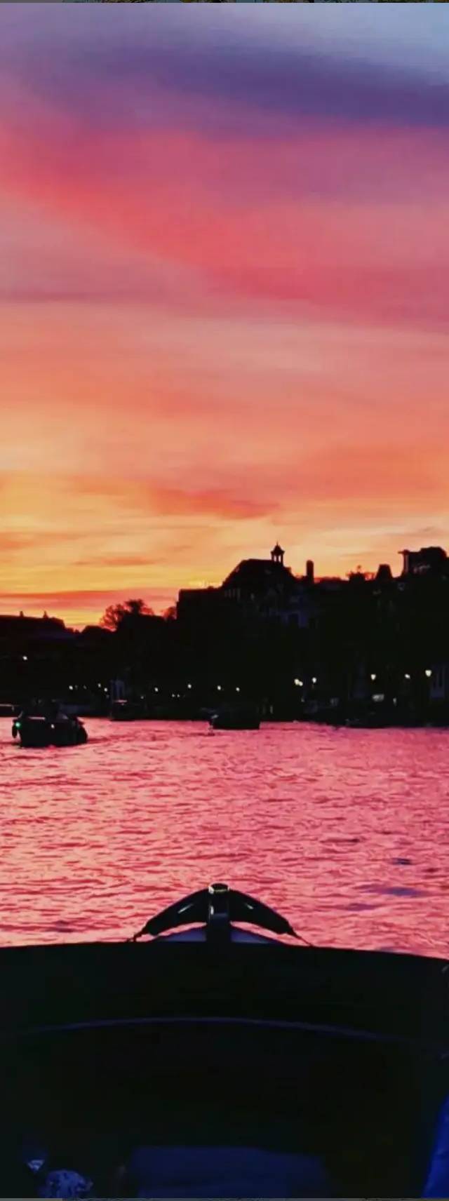 Boat bow facing a pink and orange sunset over an Amsterdam canal with silhouetted buildings