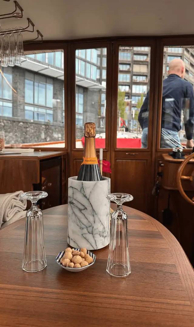 Champagne bottle in marble cooler on wooden table with two glasses and nuts, skipper at helm behind