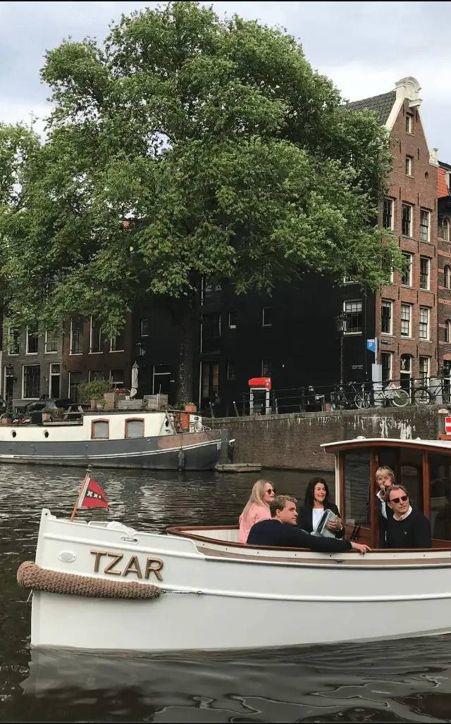 Passengers aboard the white boat Tzar on an Amsterdam canal beside brick houses and a large tree