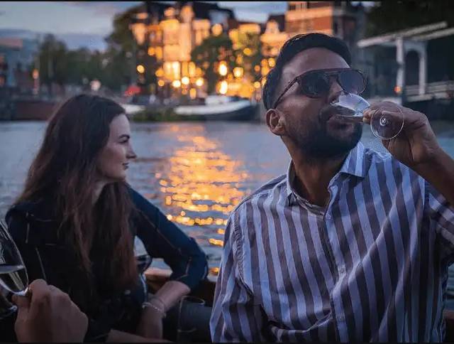 Man in sunglasses sips white wine beside a woman on an Amsterdam canal at dusk
