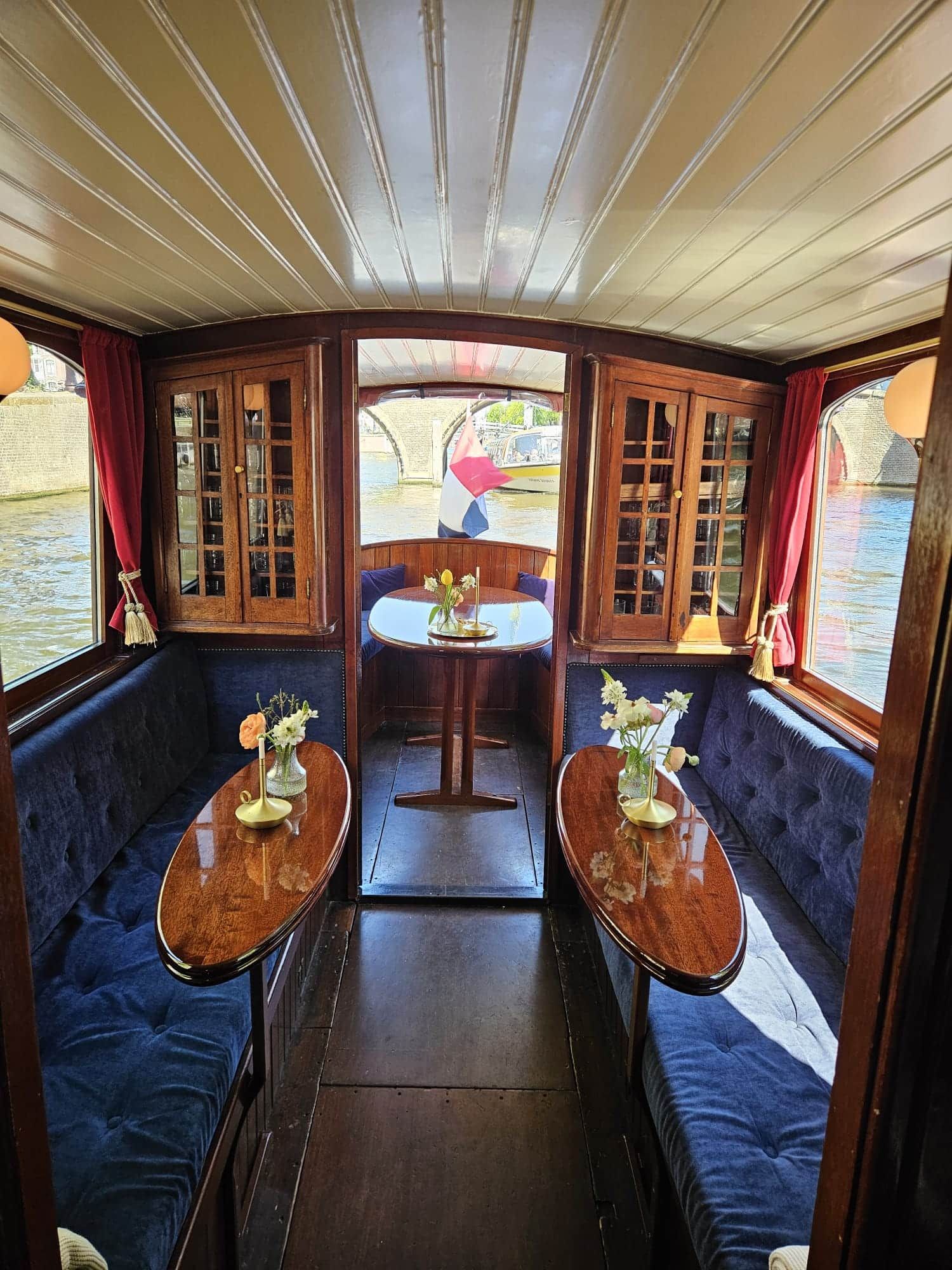 Wood-panelled saloon boat interior with flower-topped tables, aft doorway framing a Dutch flag and canal bridge