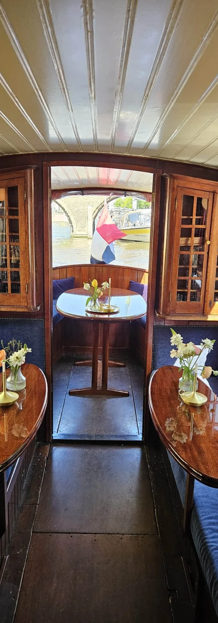 Wood-panelled saloon boat interior with flower-topped tables, aft doorway framing a Dutch flag and canal bridge