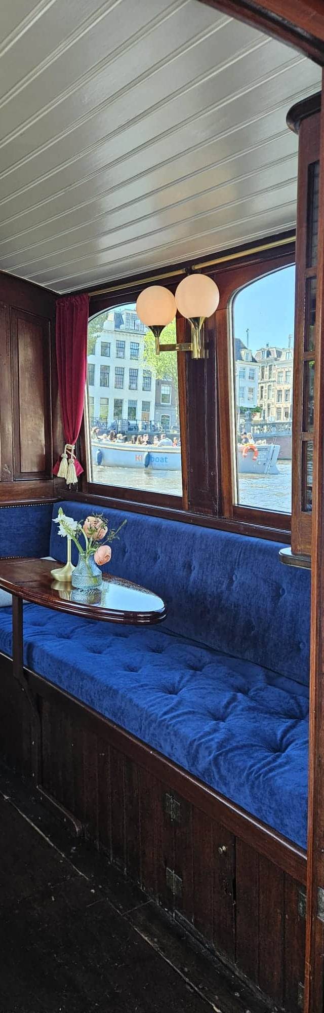 Blue velvet tufted bench and oval wooden table with flowers inside the Ritz saloon, canal houses visible through windows
