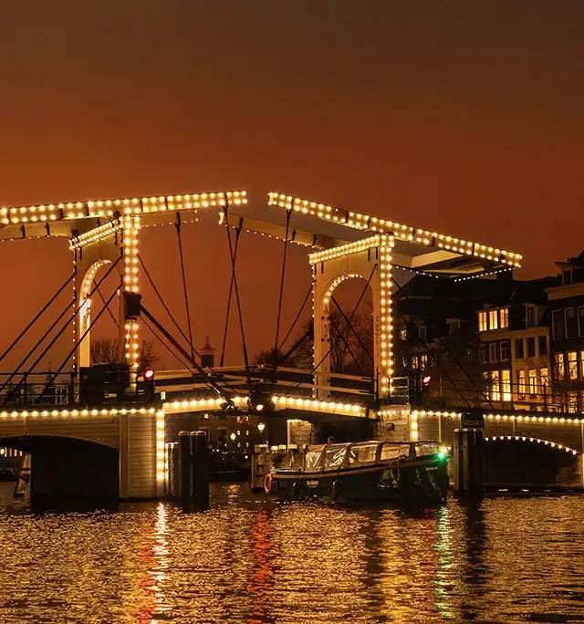 Open boat passing beneath the illuminated Magere Brug drawbridge on an Amsterdam canal at dusk