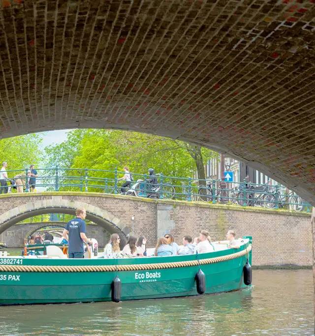 Green Eco Boats canal boat with seated passengers passing beneath a brick bridge in Amsterdam