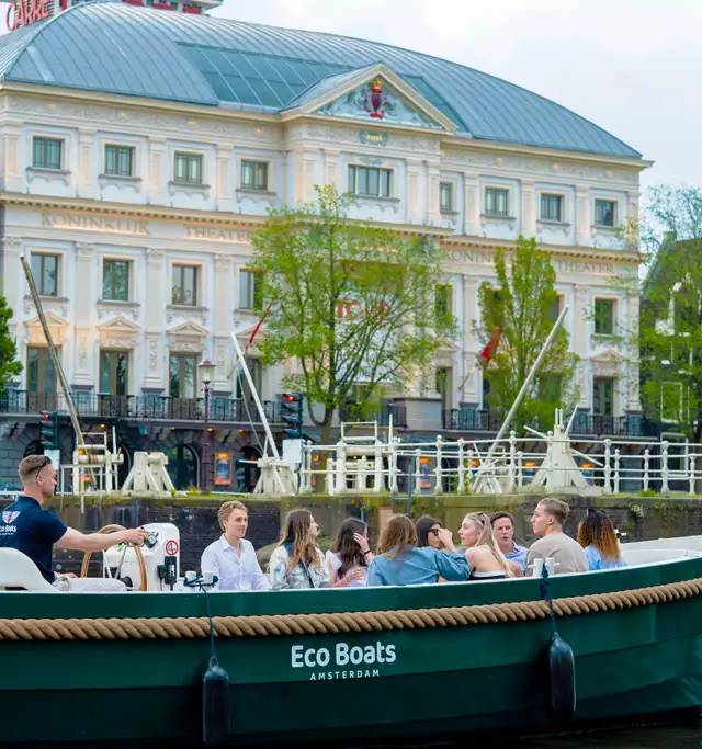 Green Eco Boats canal boat carrying a group of passengers past a white neoclassical theatre in Amsterdam