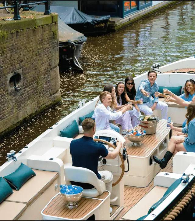 Friends raise glasses on open boat deck as a skipper steers along a brick-walled Amsterdam canal