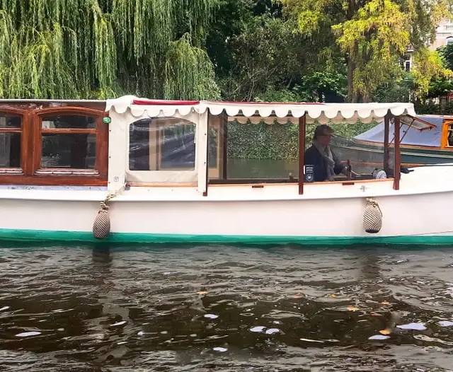 White and green wooden canal boat with scalloped canopy, helmsman at the wheel, beside a willow-lined bank