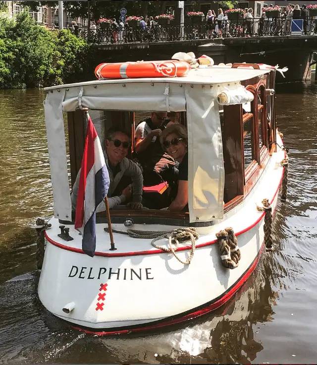 Delphine, a white and red saloon boat, carries smiling passengers under a Dutch flag on an Amsterdam canal