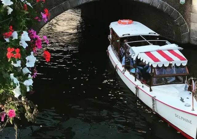 White Delphine canal boat with red-striped awning approaching a stone bridge, flower baskets in the foreground