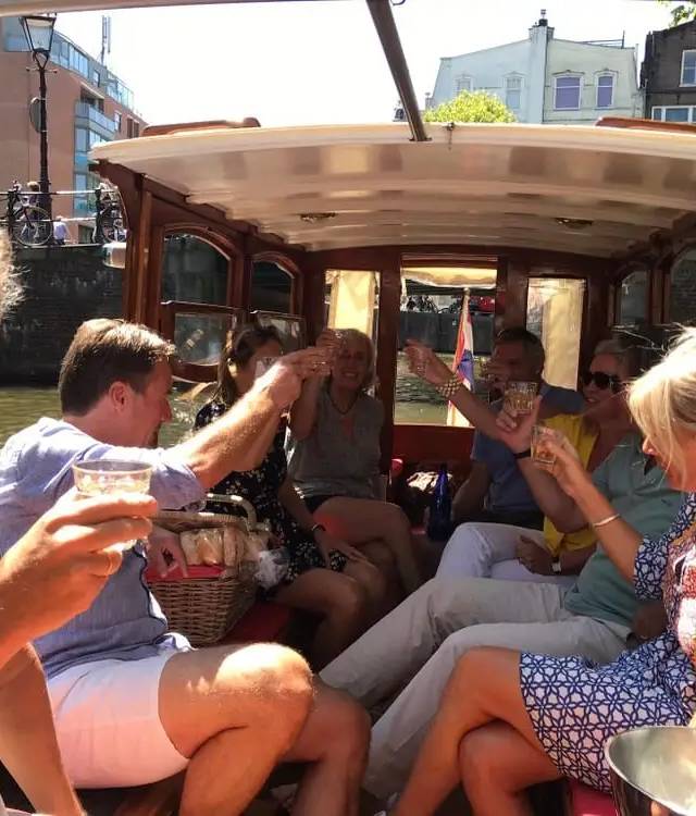 Group of friends raising glasses in a toast inside a wooden canal boat, sunny Amsterdam canal outside