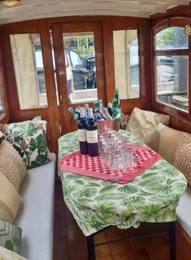 Wooden boat cabin with cushioned benches, leaf-print table holding wine bottles, glasses and an ice bucket