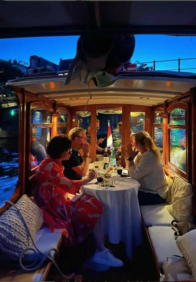 Guests seated around a white-clothed table with wine in a wooden saloon boat on an Amsterdam canal at dusk
