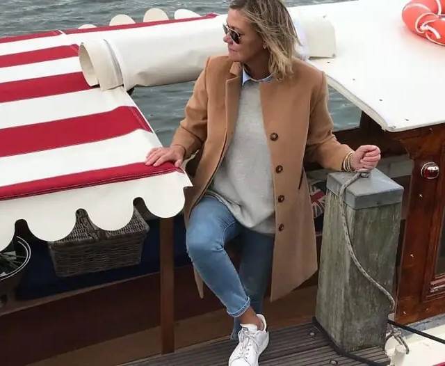 Woman in camel coat and sunglasses sits on wooden boat deck beside red-striped canopy and mooring post